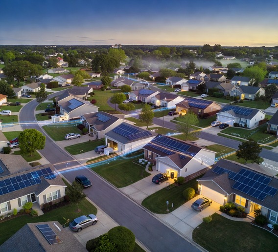 Aerial twilight view of a quiet American suburban neighborhood with glowing rooftop solar panels on dozens of homes, soft blue and gold light emanating from battery storage units visible through garage windows, interconnected by subtle glowing data lines representing a virtual power plant network, photorealistic, cinematic drone photography style, warm hopeful atmosphere, ultra high detail