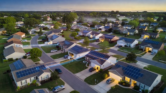 Aerial twilight view of a quiet American suburban neighborhood with glowing rooftop solar panels on dozens of homes, soft blue and gold light emanating from battery storage units visible through garage windows, interconnected by subtle glowing data lines representing a virtual power plant network, photorealistic, cinematic drone photography style, warm hopeful atmosphere, ultra high detail