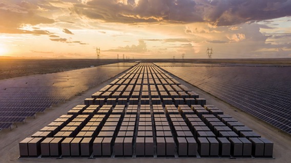Photorealistic wide-angle photograph at golden hour of a massive Tesla Megapack battery installation stretching to the horizon, hundreds of sleek graphite-colored rectangular units arranged in precise geometric rows, flanked on both sides by vast solar panel arrays catching the last warm light of sunset, dramatic clouds above, electric transmission towers visible in the distance, conveying industrial scale and clean energy optimism, cinematic lighting, ultra-detailed, 8K resolution
