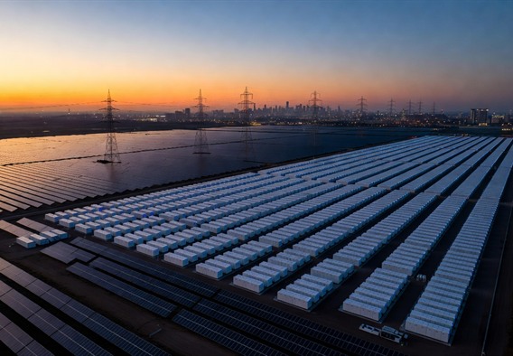 Sweeping aerial drone photograph at twilight of a massive Tesla Megapack battery installation arranged in precise geometric rows, glowing with soft blue indicator lights, adjacent to a vast solar farm with panels catching the last orange light of sunset, high-voltage transmission towers visible in the background connecting to a distant city skyline, photorealistic, cinematic lighting, ultra-high detail, sense of enormous scale and clean energy abundance