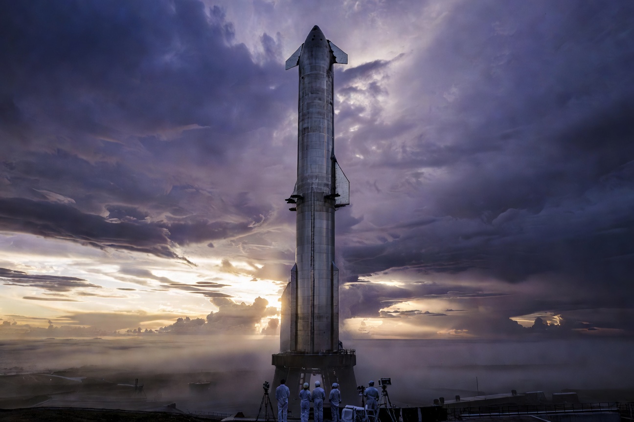 Starship rocket on the launchpad at Boca Chica with a dramatic stormy sky overhead, symbolizing the tension between ambition and physical reality