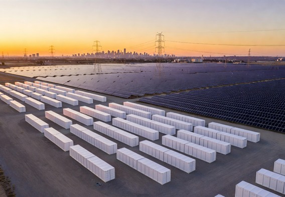 Aerial drone photograph at golden hour of a massive Tesla Megapack installation comprising hundreds of white rectangular battery units arranged in precise geometric rows, immediately adjacent to a sprawling solar farm with thousands of dark blue photovoltaic panels, soft amber light reflecting off both surfaces, power transmission lines visible in the background connecting to distant city lights on the horizon, ultra-realistic photographic style, cinematic lighting, 8k resolution
